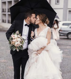 Gorgeous bride and stylish groom walking under umbrella in rainy street and kissing. Sensual wedding couple embracing. Romantic moments of newlyweds. Modern  wedding photo